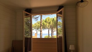 A bedroom with a large window, a bed, and a nightstand with a lamp. - Résidence Les Pins (Calvi)