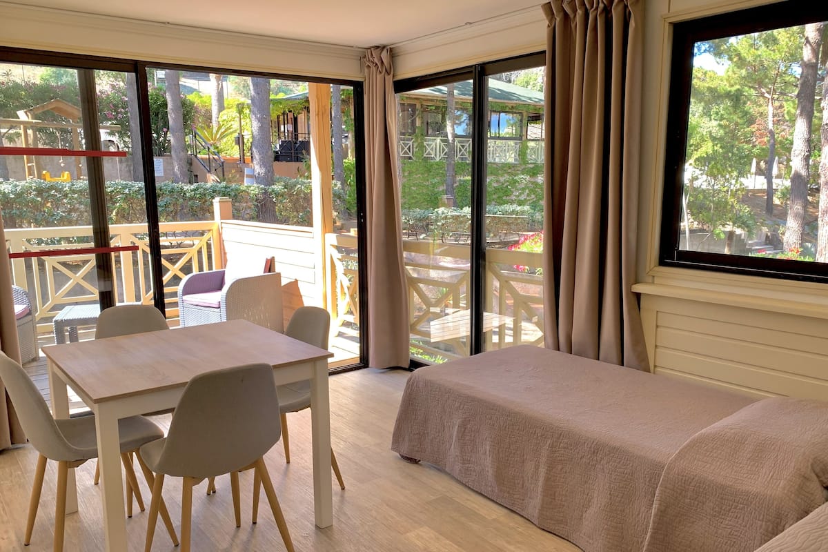 A room with a wooden table and chairs, a bed, and large windows offering a view of a garden and outdoor seating area.