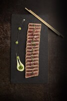A black slate serving board with thinly sliced meat arranged in a row, garnished with small flowers and green beads, accompanied by chopsticks and a small white spoon with green beads.