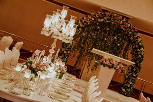 Una mesa de banquete dispuesta con cristalería y centros de flores, una araña y un arco floral.
