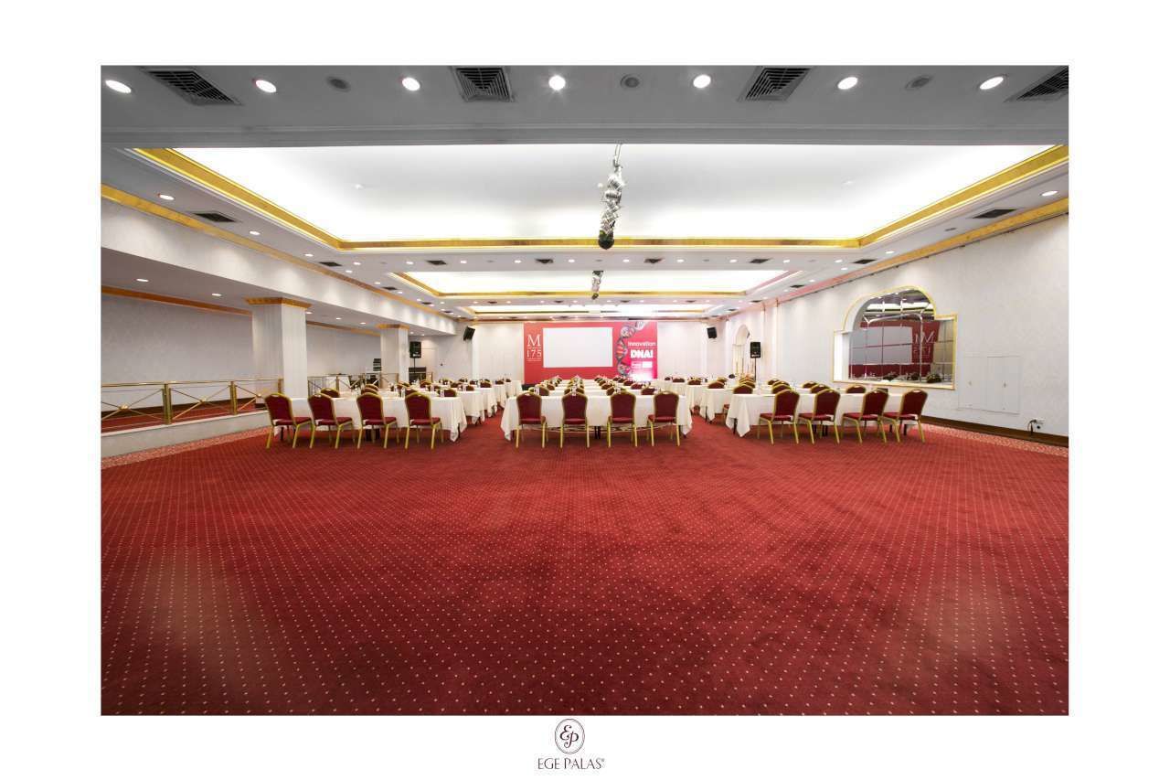 Un amplio salón de conferencias con filas de mesas y sillas, suelo cubierto de moqueta roja y un escenario con pantalla.