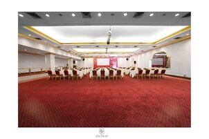 Un amplio salón de conferencias con filas de mesas y sillas, suelo cubierto de moqueta roja y un escenario con pantalla.
