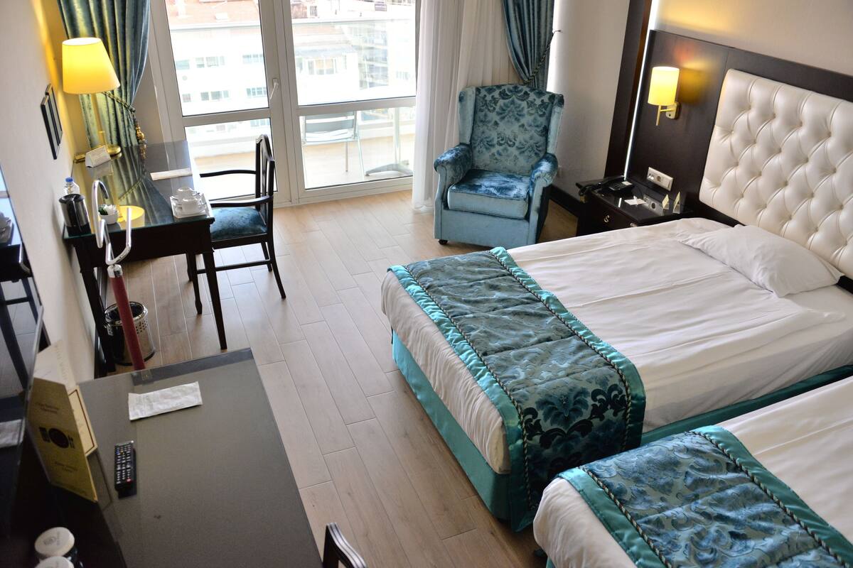 A hotel room with two beds, a desk with a chair, a lamp, and a chair by the window.