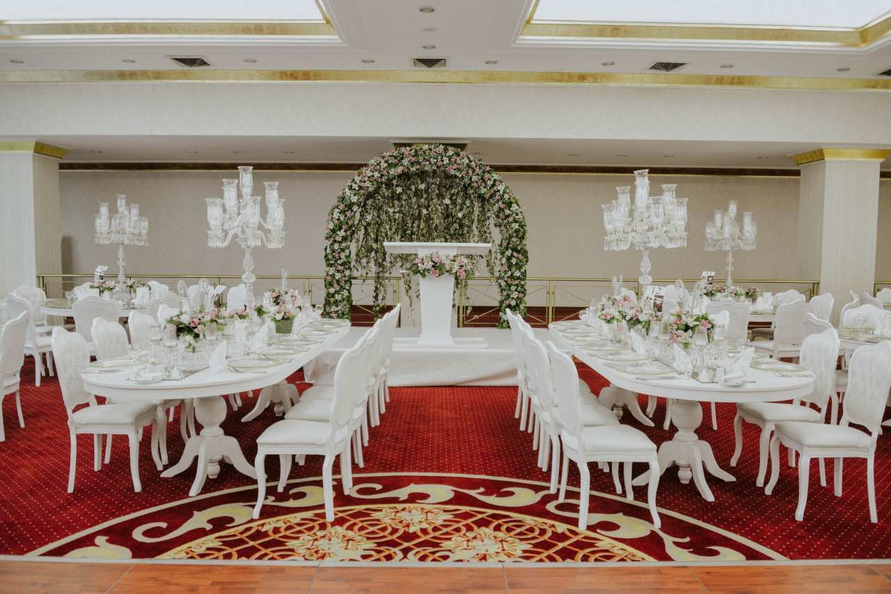 Un salón de banquetes con mesas redondas, sillas blancas y un arco floral.