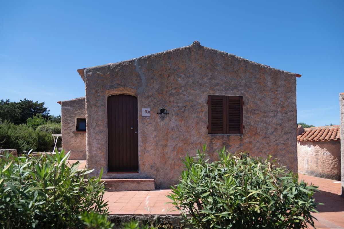 A stone building with a wooden door and window, surrounded by greenery.