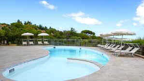 A swimming pool with a curved edge, surrounded by lounge chairs and umbrellas, with a view of trees and hills in the background. - AHR Altura Hotel Porto Rafael (Palau)