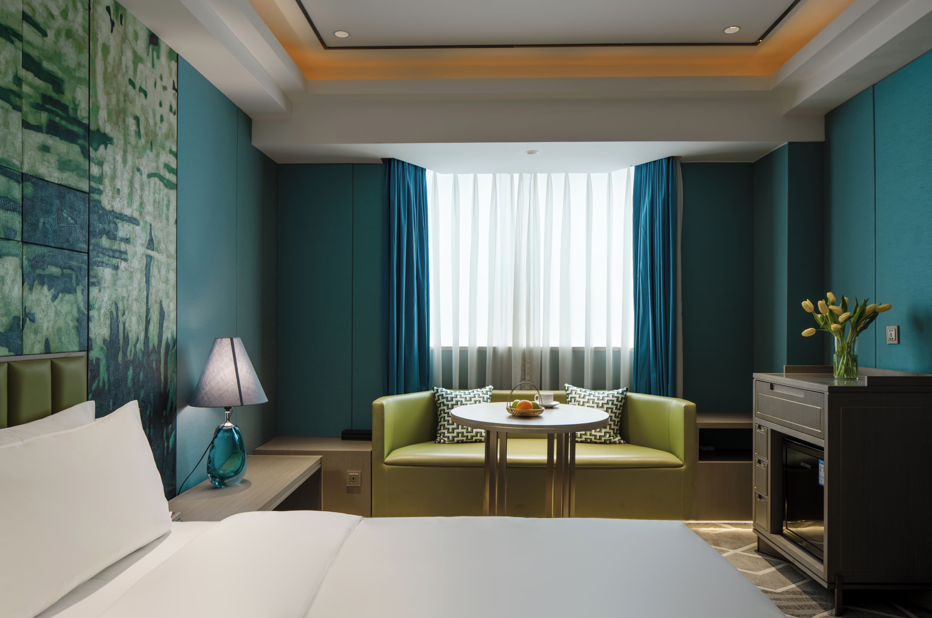 A hotel room with a bed, a green sofa, a small table with a bowl, a lamp, a painting on the wall, and a window with curtains.