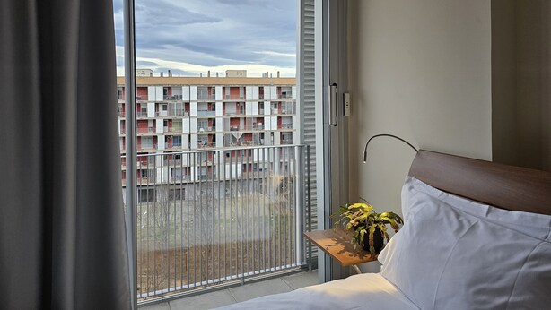 Habitación de hotel con puerta corrediza de vidrio a un balcón, una pequeña mesita de madera con una planta y una cama con ropa blanca.