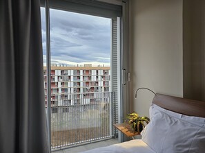 A hotel room with a sliding glass door to a balcony, a small wooden side table with a plant, and a bed with white bedding.