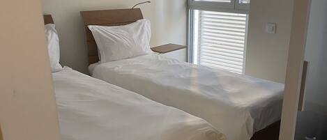 A bedroom with two single beds, a window with blinds, and a door leading to another room.