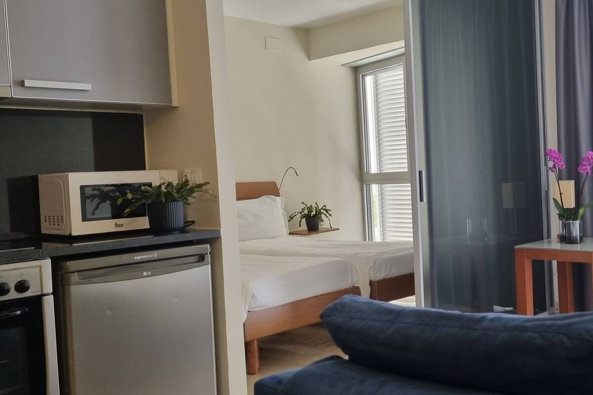 Habitación de hotel con cama, una mesita con un florero, televisión y una kitchenette con microondas y refrigerador.