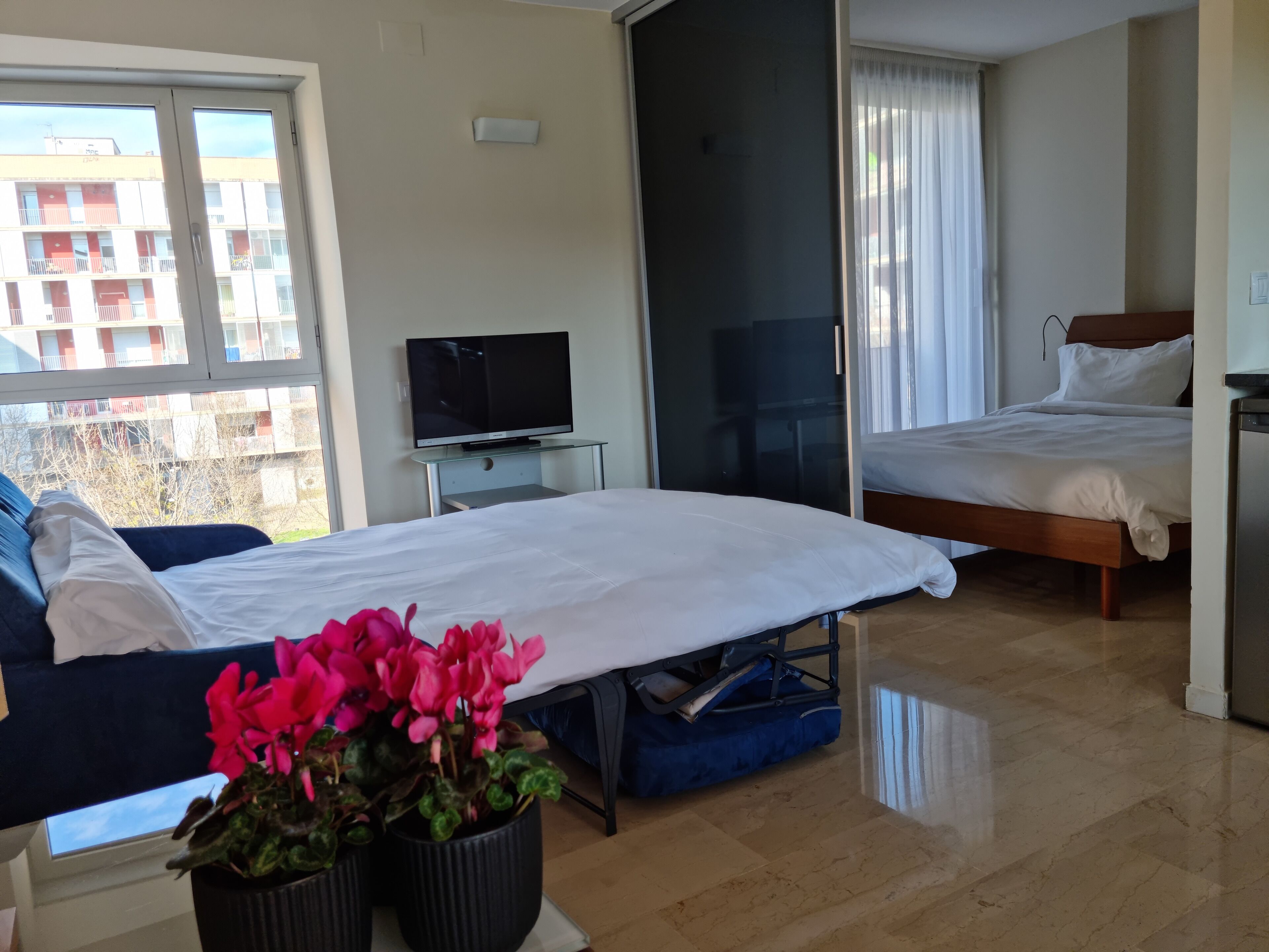 A hotel room with a bed, a TV, a sliding door, and a view of buildings outside.