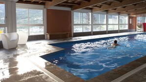 A swimming pool with a person swimming, large windows, and a wooden ceiling. - Le Château des Magnans (Jausiers)