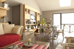 A living room with a sofa, a coffee table with magazines, a bookshelf, and a dining area with chairs and a table. - Le Château des Magnans (Jausiers)