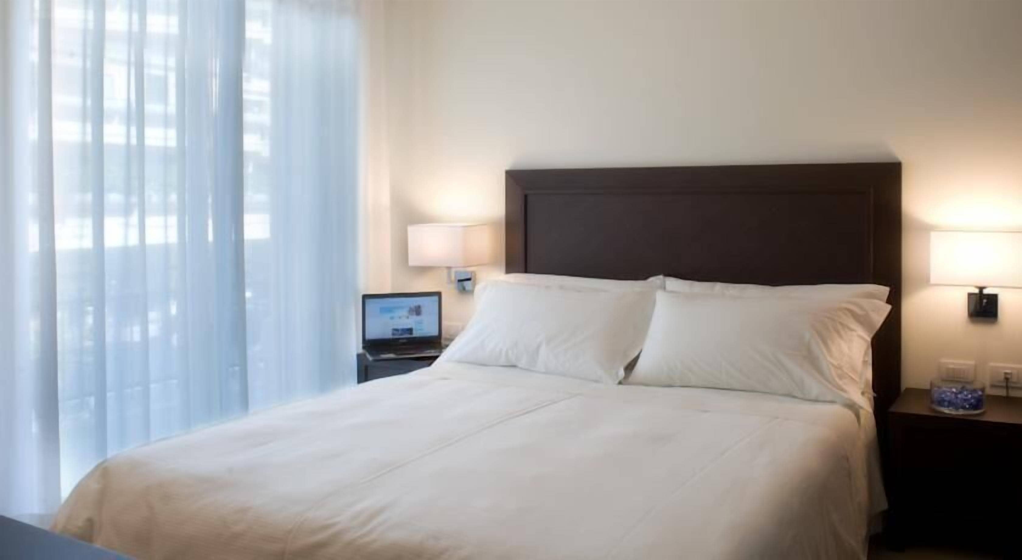Habitación de hotel con una cama grande, dos lámparas de noche, un ordenador portátil sobre una mesita y una ventana con cortinas traslúcidas.