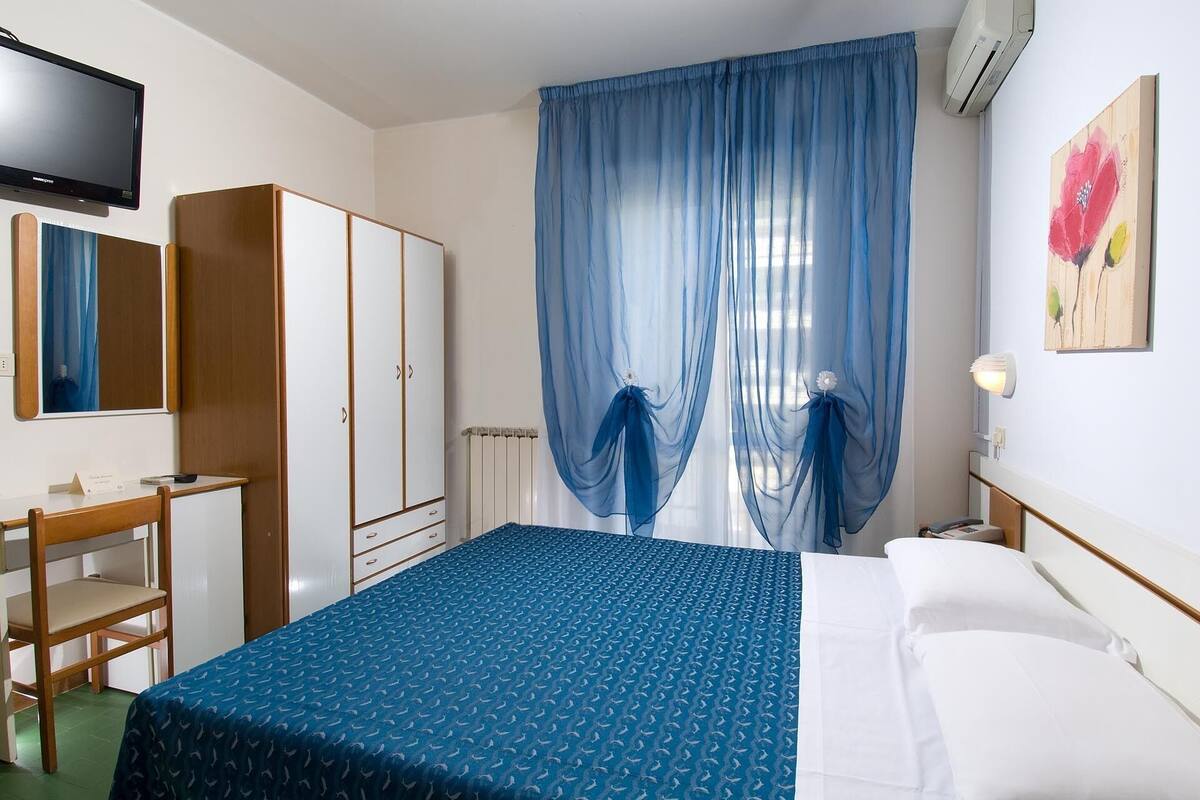 A hotel room with a bed, a desk with a chair, a wardrobe, a radiator, and a painting on the wall.