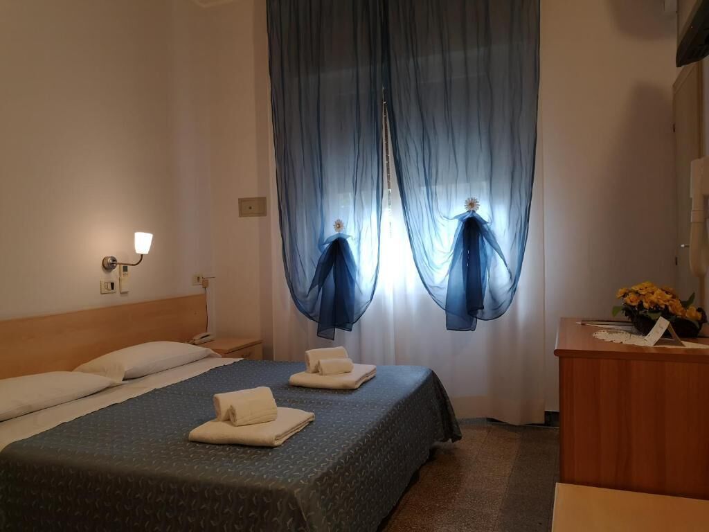 A hotel room with a bed, a nightstand, a desk with a lamp, and sheer curtains.