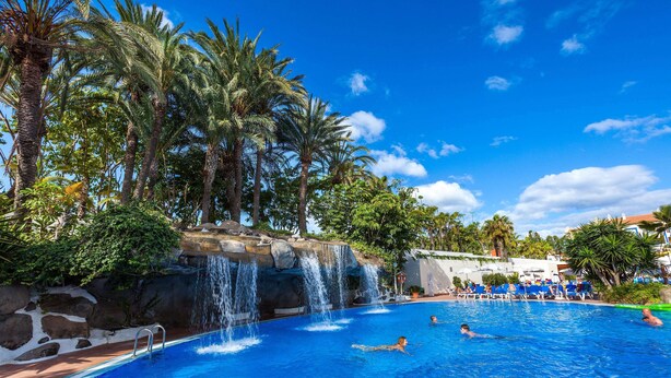 A swimming pool with a waterfall feature, surrounded by palm trees and a clear blue sky. - Hotel Best Tenerife (Arona)