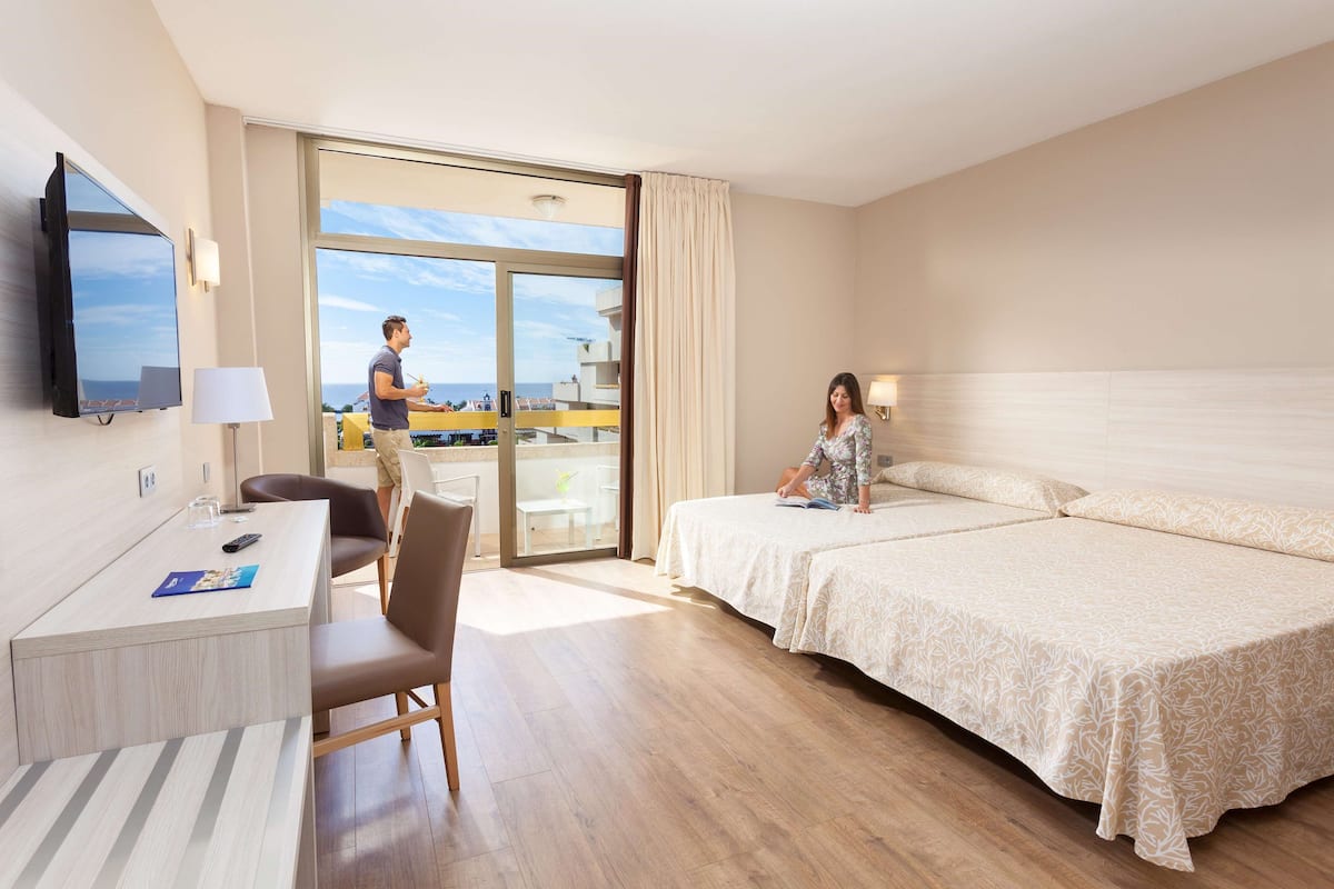 A hotel room with a bed, a desk, a chair, a TV, and a view of the sea.