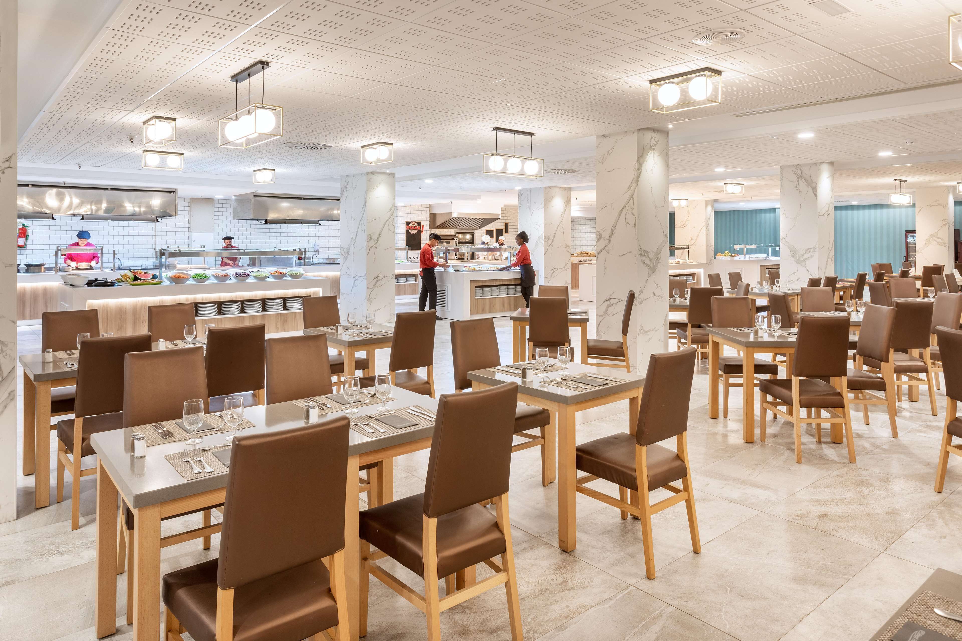 Uma sala de jantar espaçosa com várias mesas e cadeiras, um ponto central de serviço e uma cozinha ao fundo.