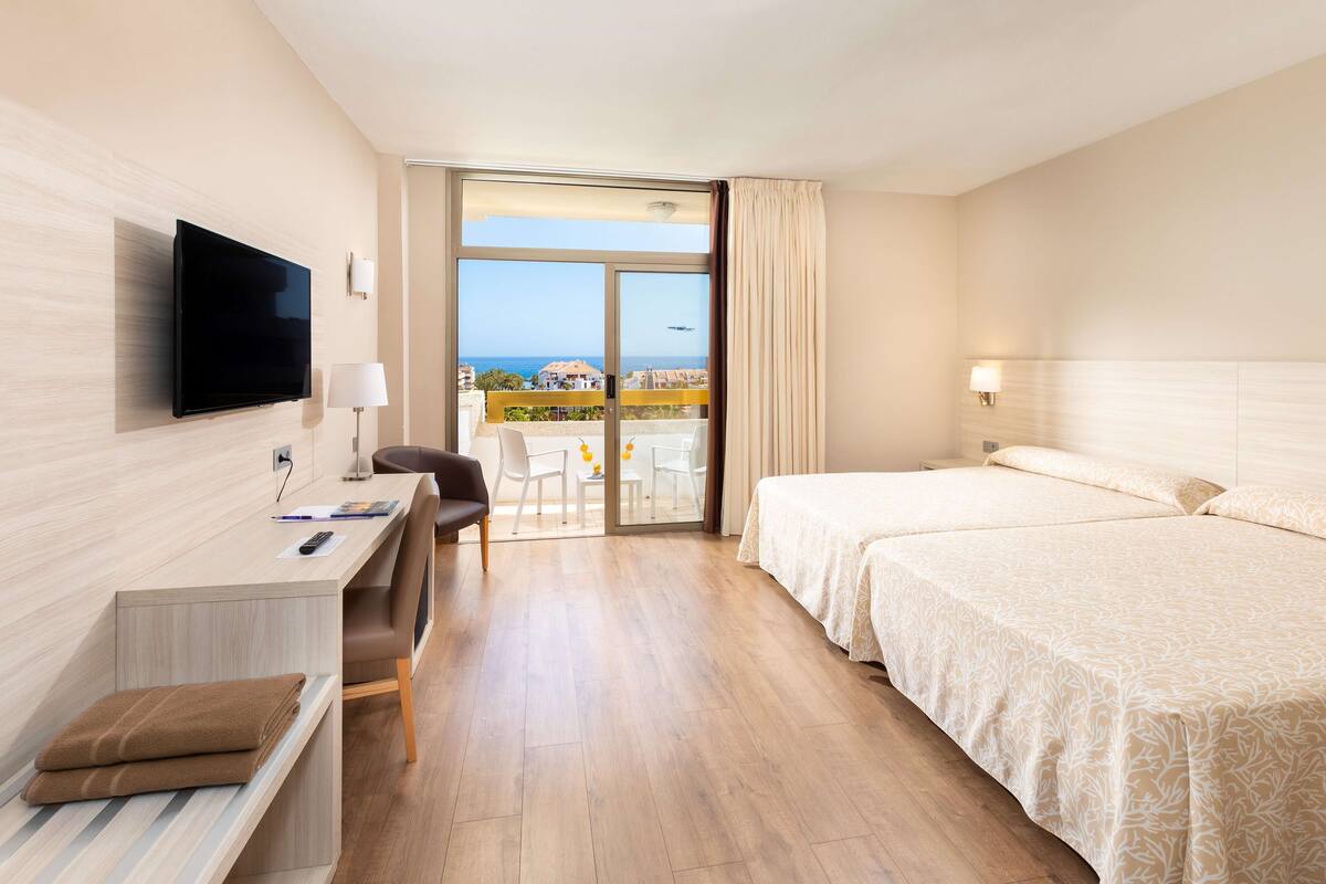 A hotel room with a desk, two beds, a TV, and a balcony with a view.