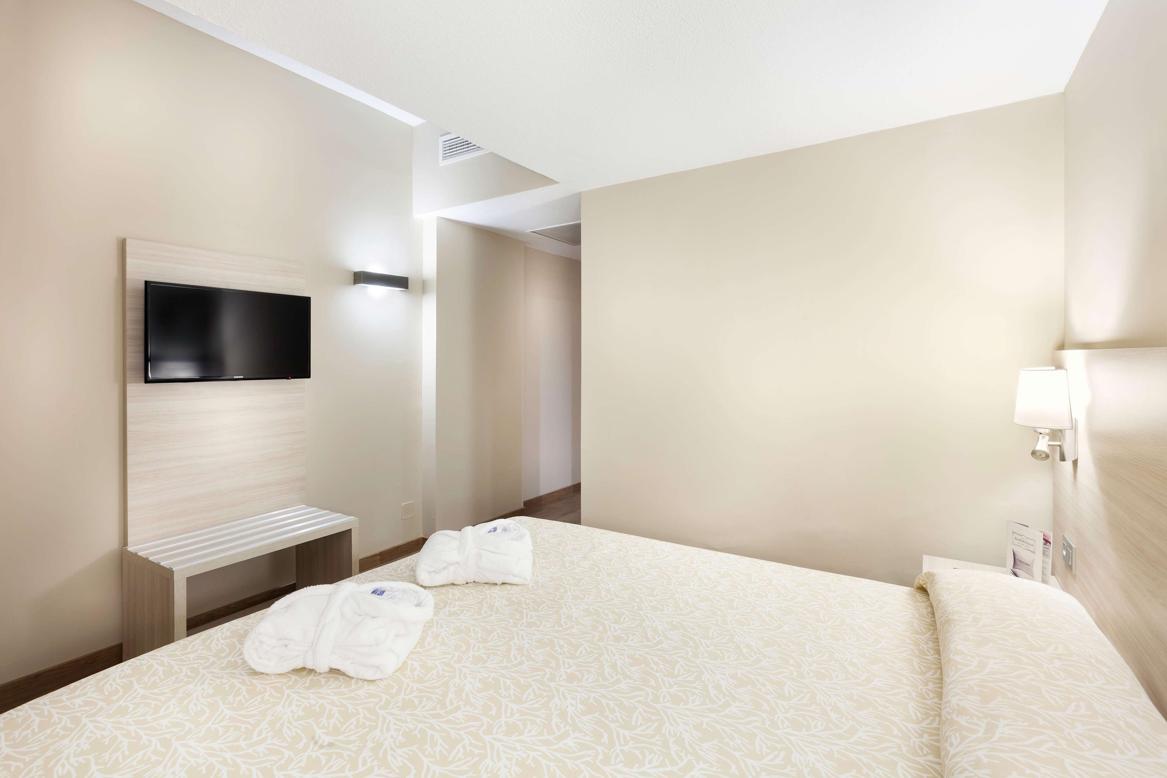 A hotel room with a bed, a TV mounted on the wall, a bench, and a pair of towels on the bed.
