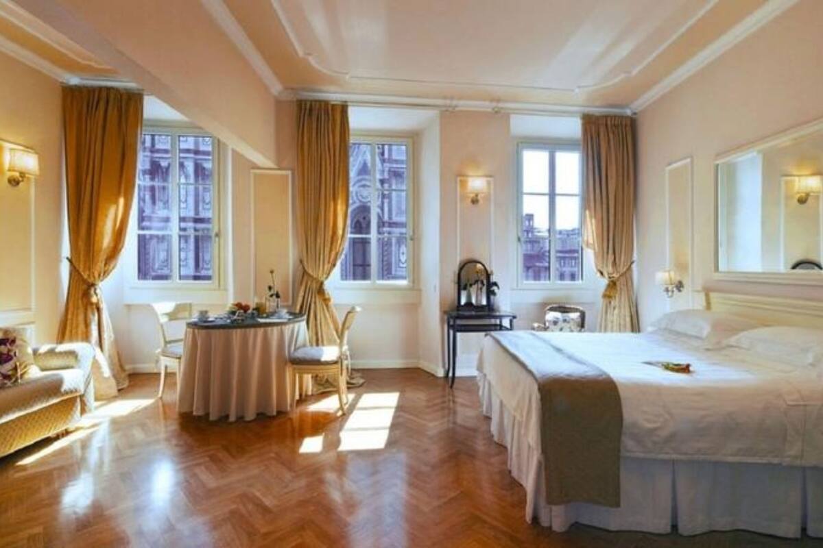 Habitación de hotel con cama, sofá, mesa de comedor y una silla. Hay ventanas con cortinas y un cuadro en la pared.