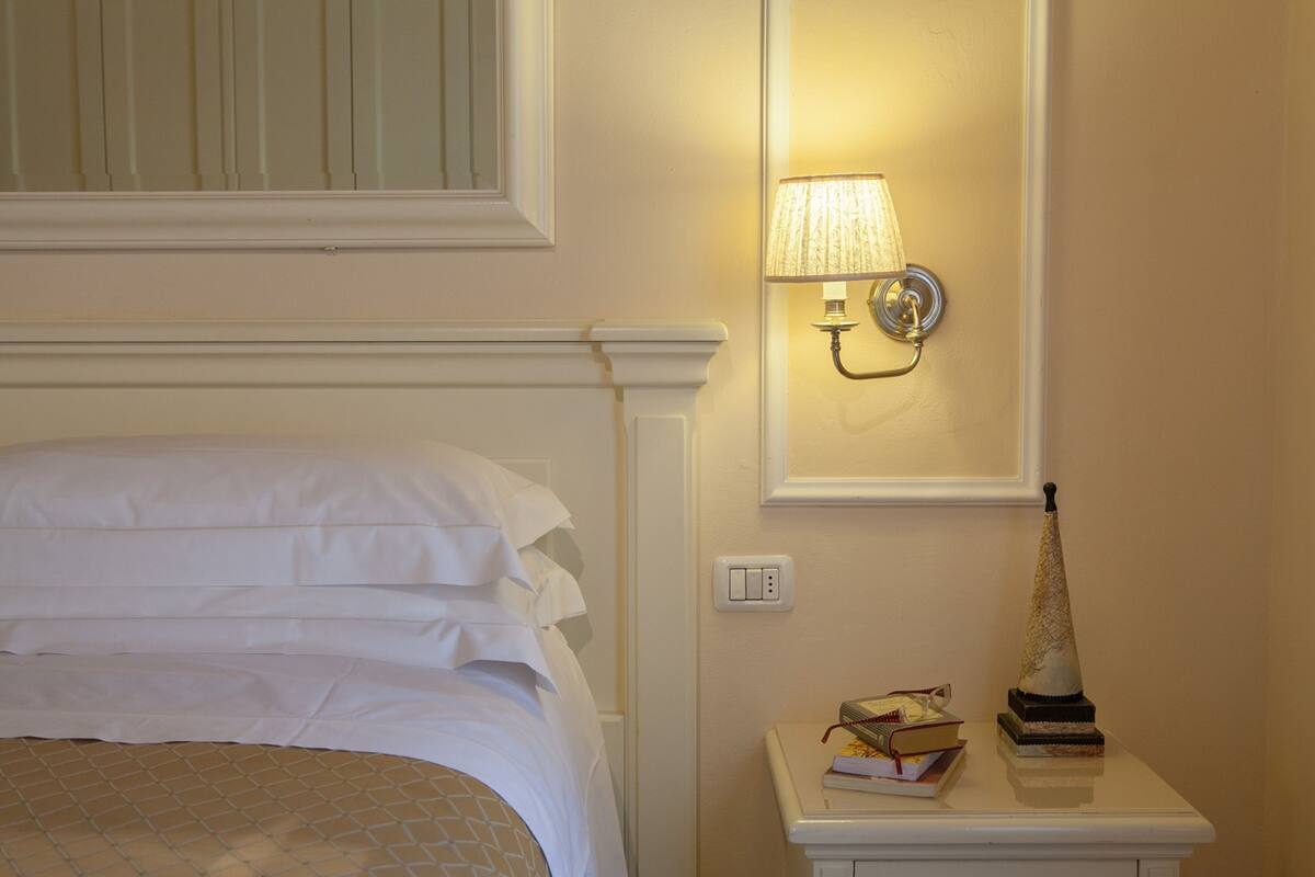 Una cama bien hecha con ropa de cama blanca, una lámpara de noche, una mesita con un libro y un objeto decorativo, y una lámpara fijada en la pared.