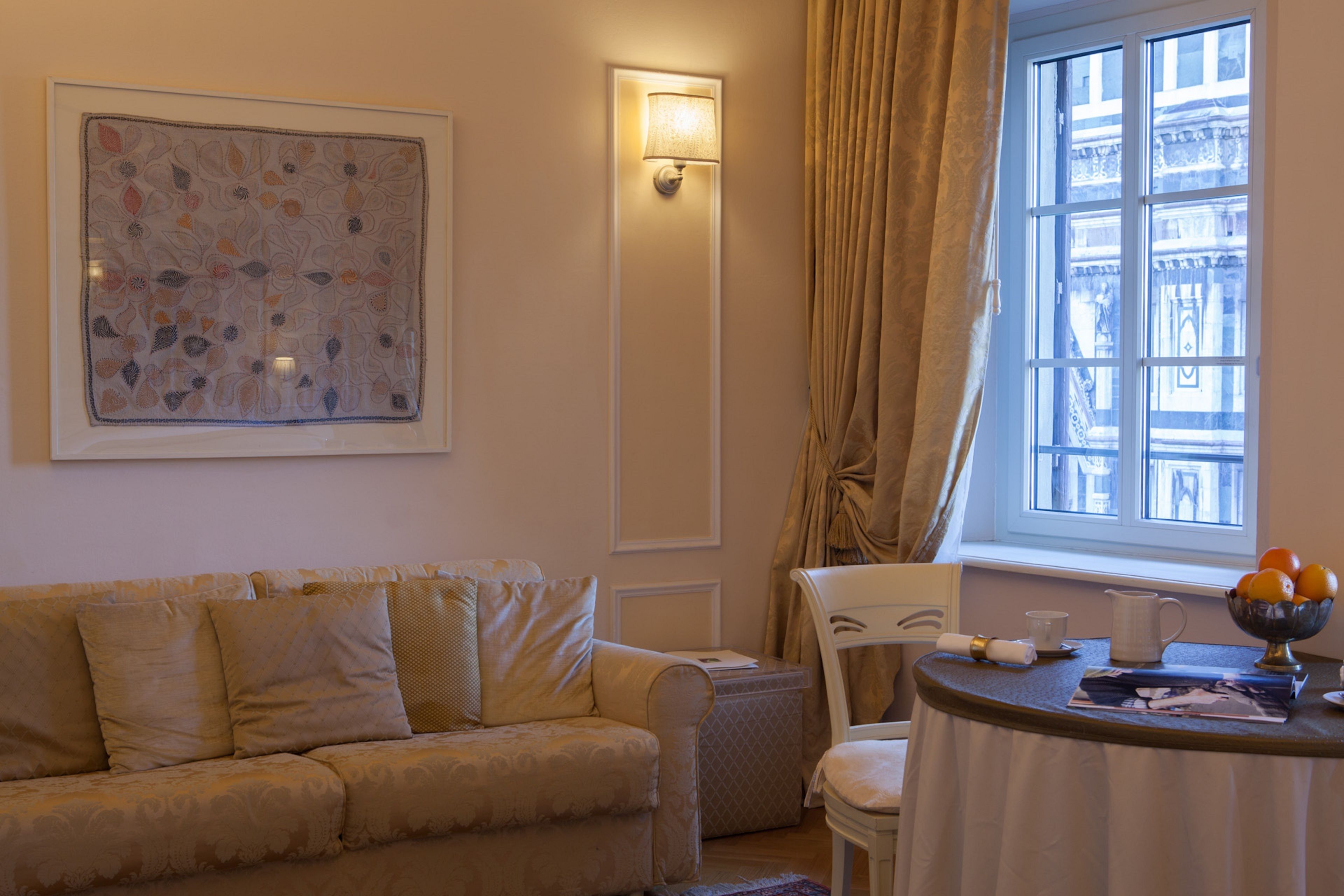 A living room with a beige sofa, a white armchair, a table with a bowl of fruit, and a window with curtains.