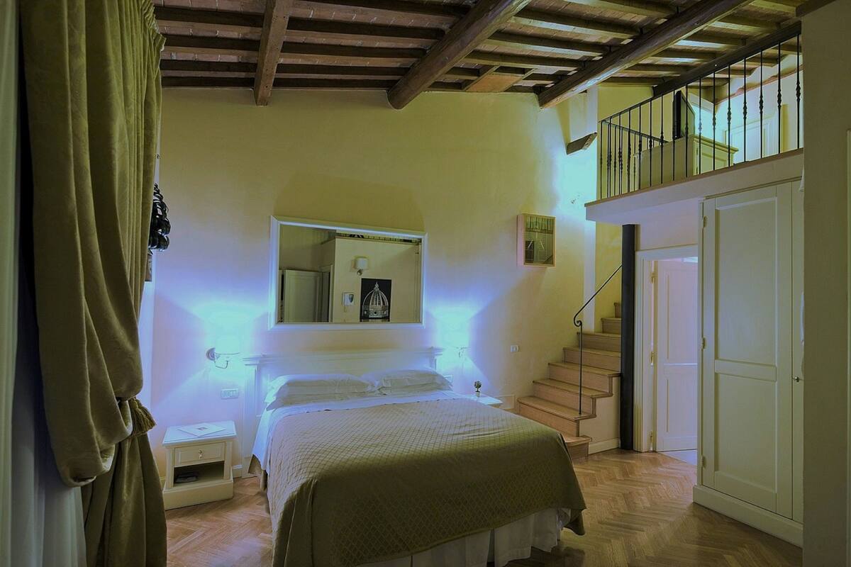 Un dormitorio con cama, mesita de noche y espejo encima. Se ve una escalera que sube a un nivel superior.