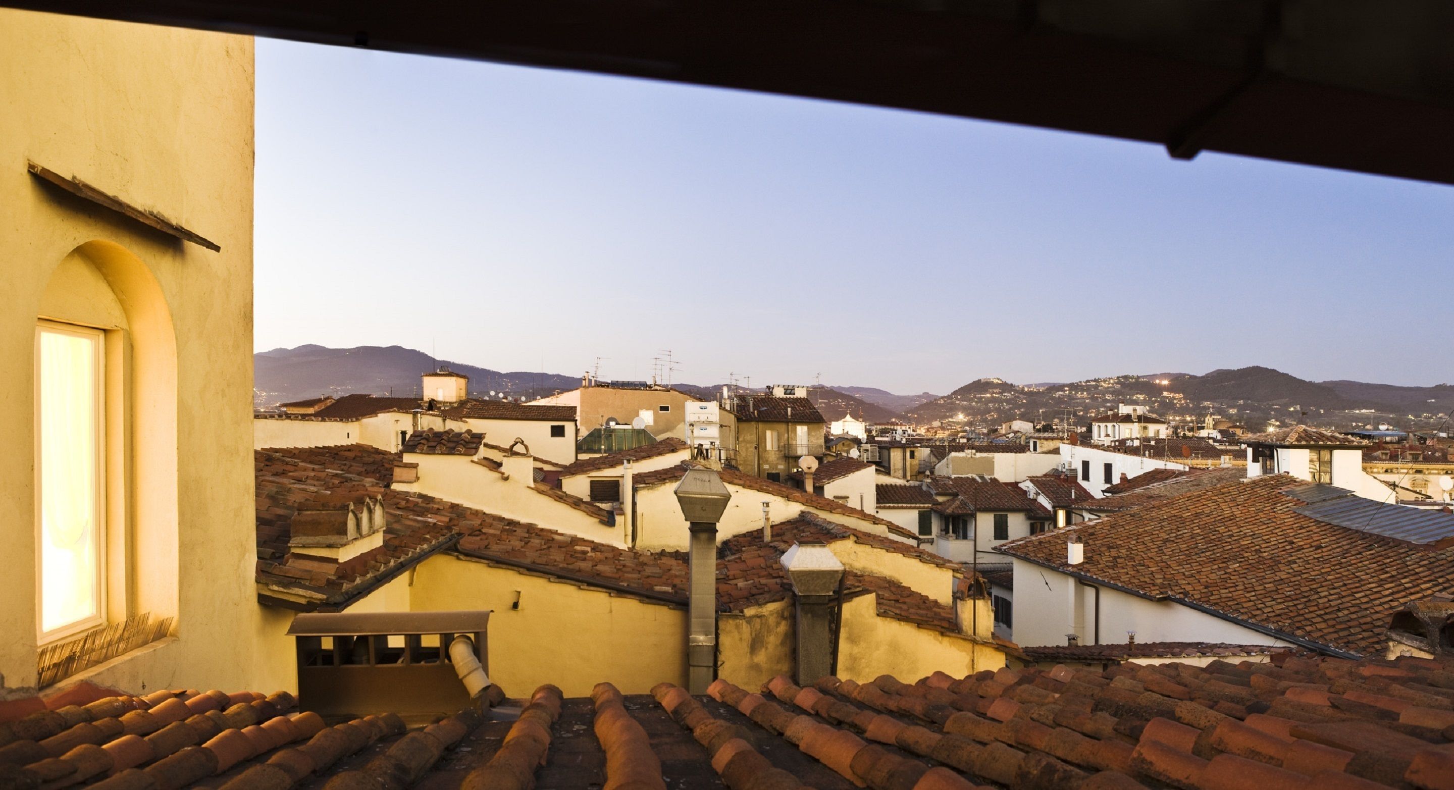 Vista de un paisaje urbano con tejados de terracota, edificios de diferentes alturas y montañas distantes bajo un cielo despejado.