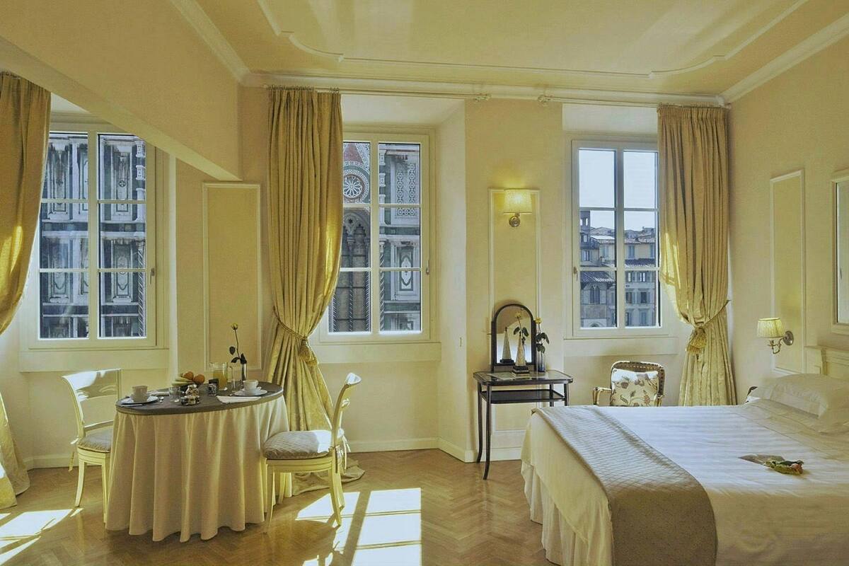 Una habitación de hotel con una cama, una mesa redonda de comedor, una silla, una mesita de noche con espejo y una ventana con cortinas.