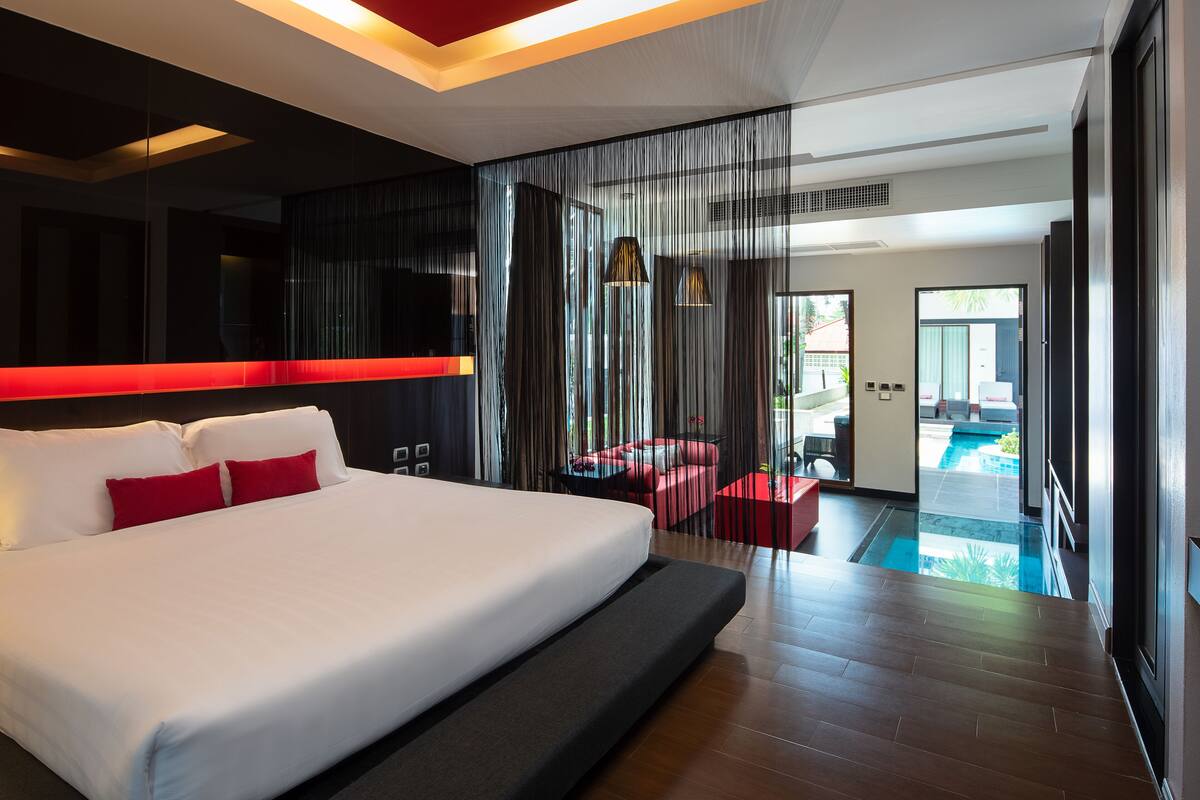 Quarto de hotel moderno com uma cama grande, uma parede de destaque vermelha, vista para uma piscina e varanda com área de estar ao ar livre.