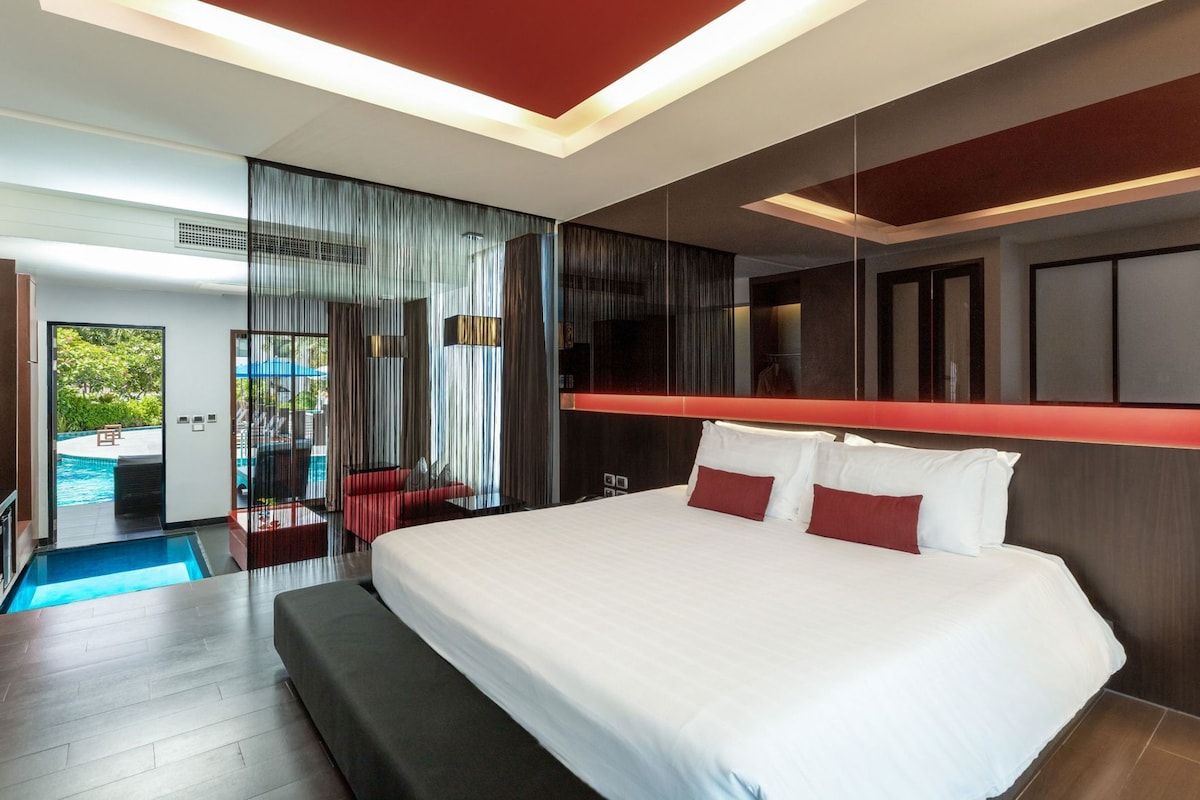 A modern hotel room with a large bed, a red accent wall, and a view of a swimming pool.