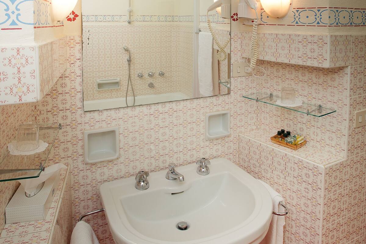 A bathroom with a sink, mirror, and shower. There are shelves with toiletries and a towel rack.