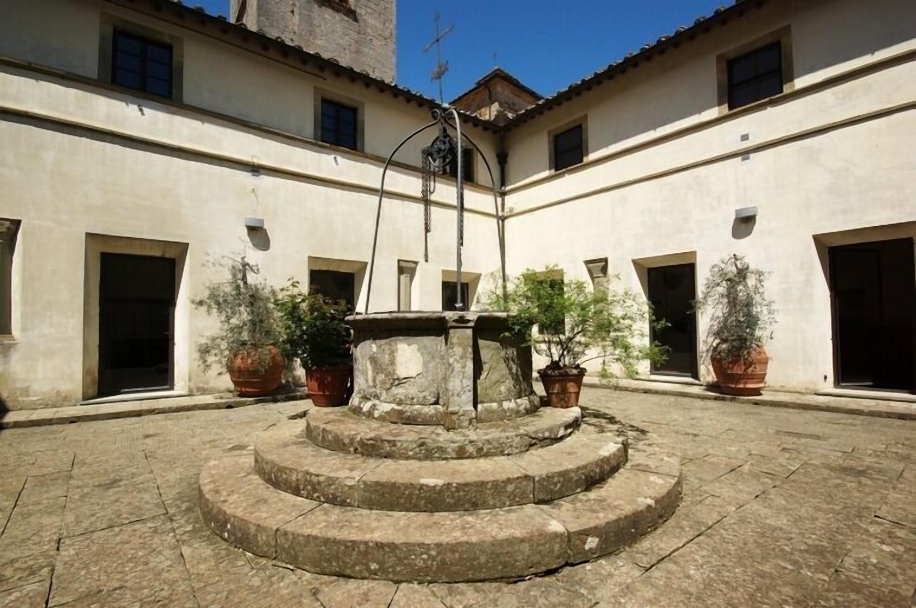 Un cortile con una fontana al centro, circondato da piante in vaso e edifici con porte e finestre.