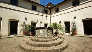 Un cortile con una fontana al centro, circondato da piante in vaso e edifici con porte e finestre.