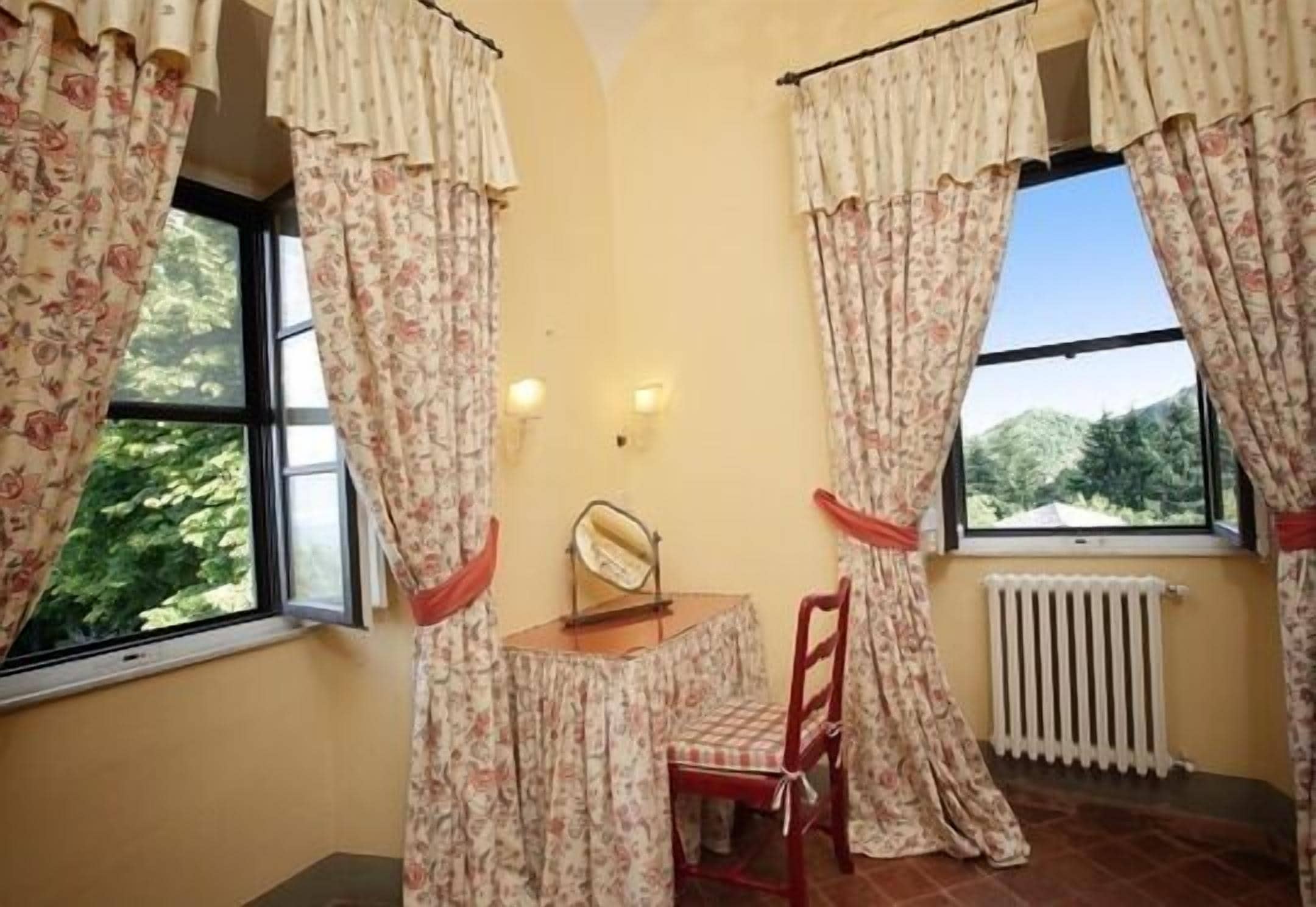 Una stanza con due finestre, un termosifone, un tavolo e una sedia, e tende.