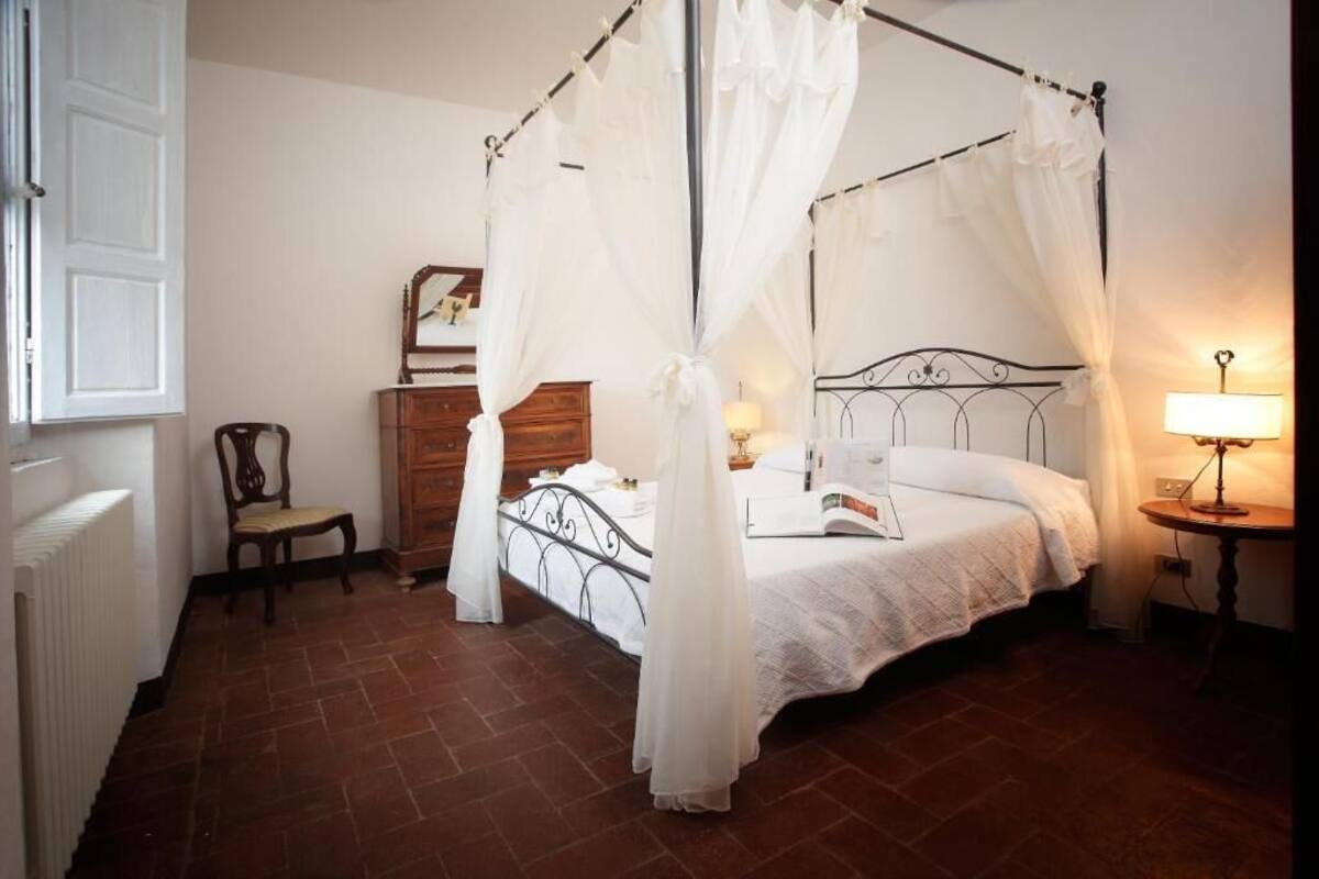 A four-poster canopy bed with a white bedspread and pillows, a wooden dresser, a chair, and a bedside table with a lamp.