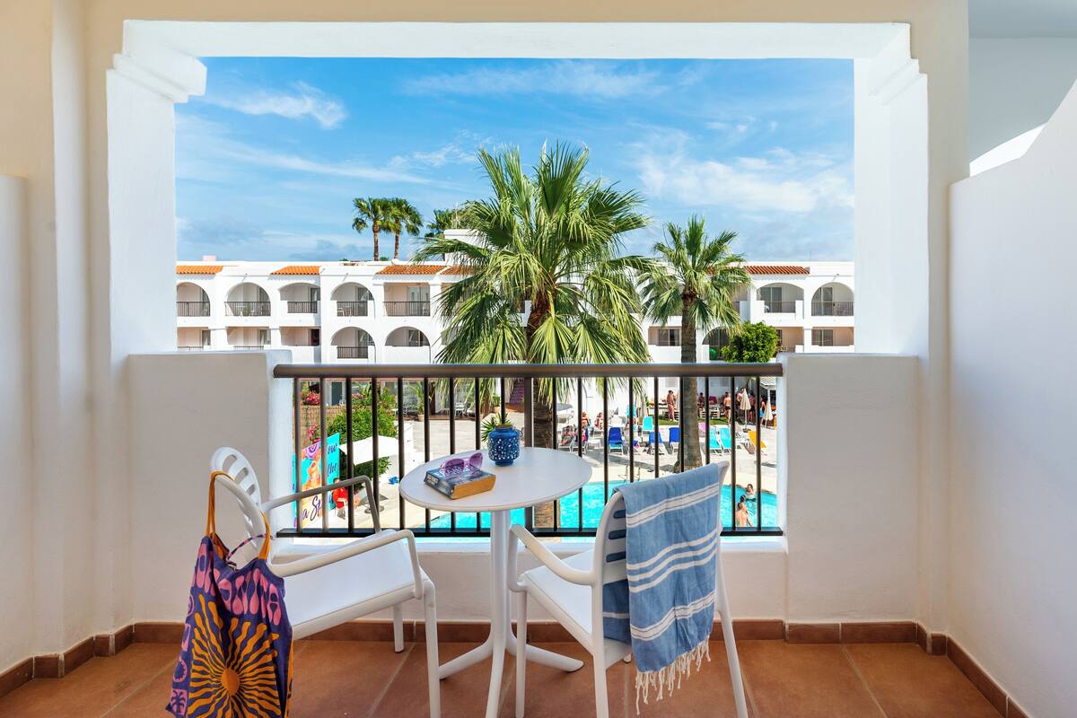 A balcony with white chairs, a small table, and a view of a pool and buildings.