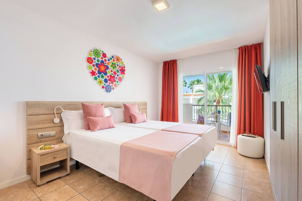 A double bed with a wooden headboard, a bedside table with a book, a decorative heart on the wall, and a balcony with red curtains.