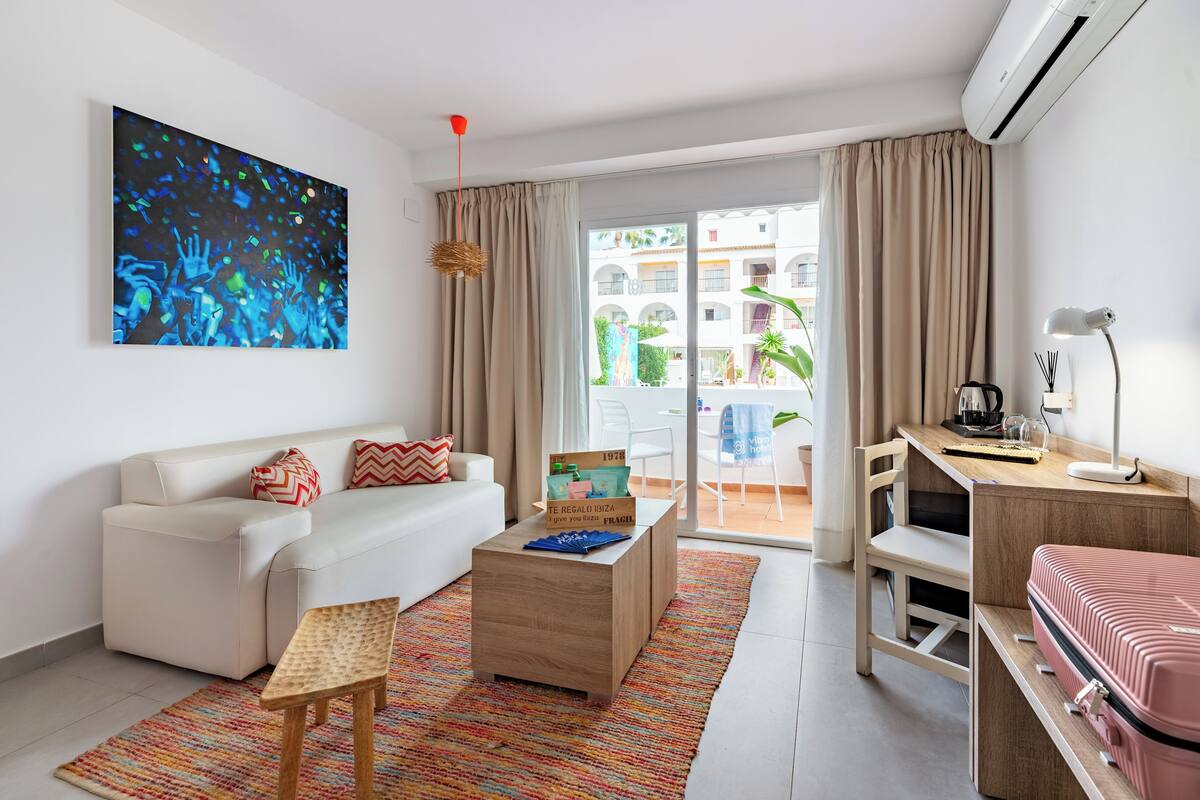 A modern hotel room with a white sofa, a wooden coffee table, a desk with a chair, and a large window with curtains.