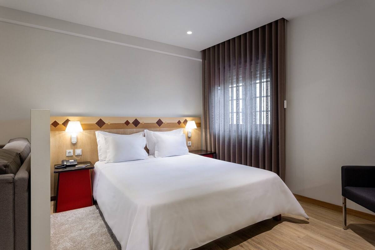 Ein Hotelzimmer mit einem großen Bett, einem Nachttisch, einem Sessel und einem Fenster mit Vorhängen.