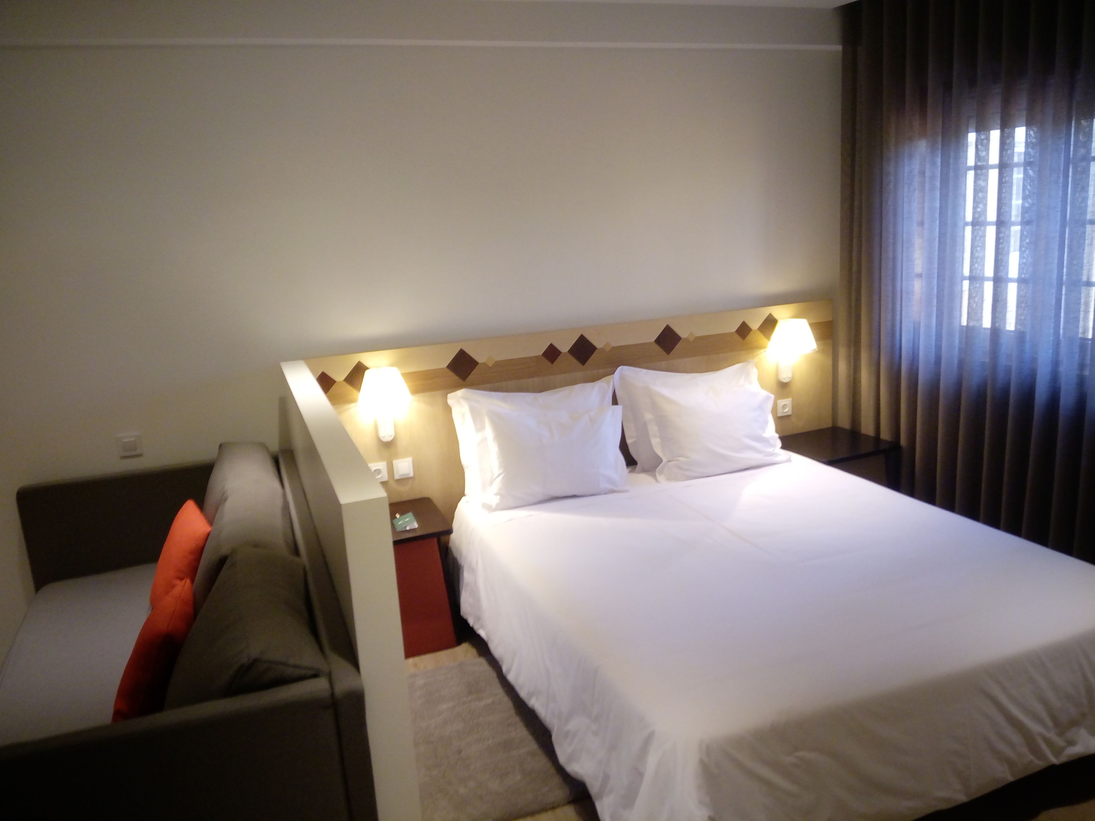 A hotel room with a bed, two bedside lamps, a nightstand, a chair, and a window with curtains.