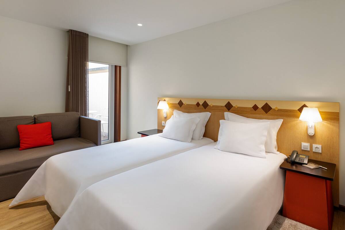 Ein Hotelzimmer mit zwei Betten, einem Schlafsofa und einem Fenster mit Vorhängen.