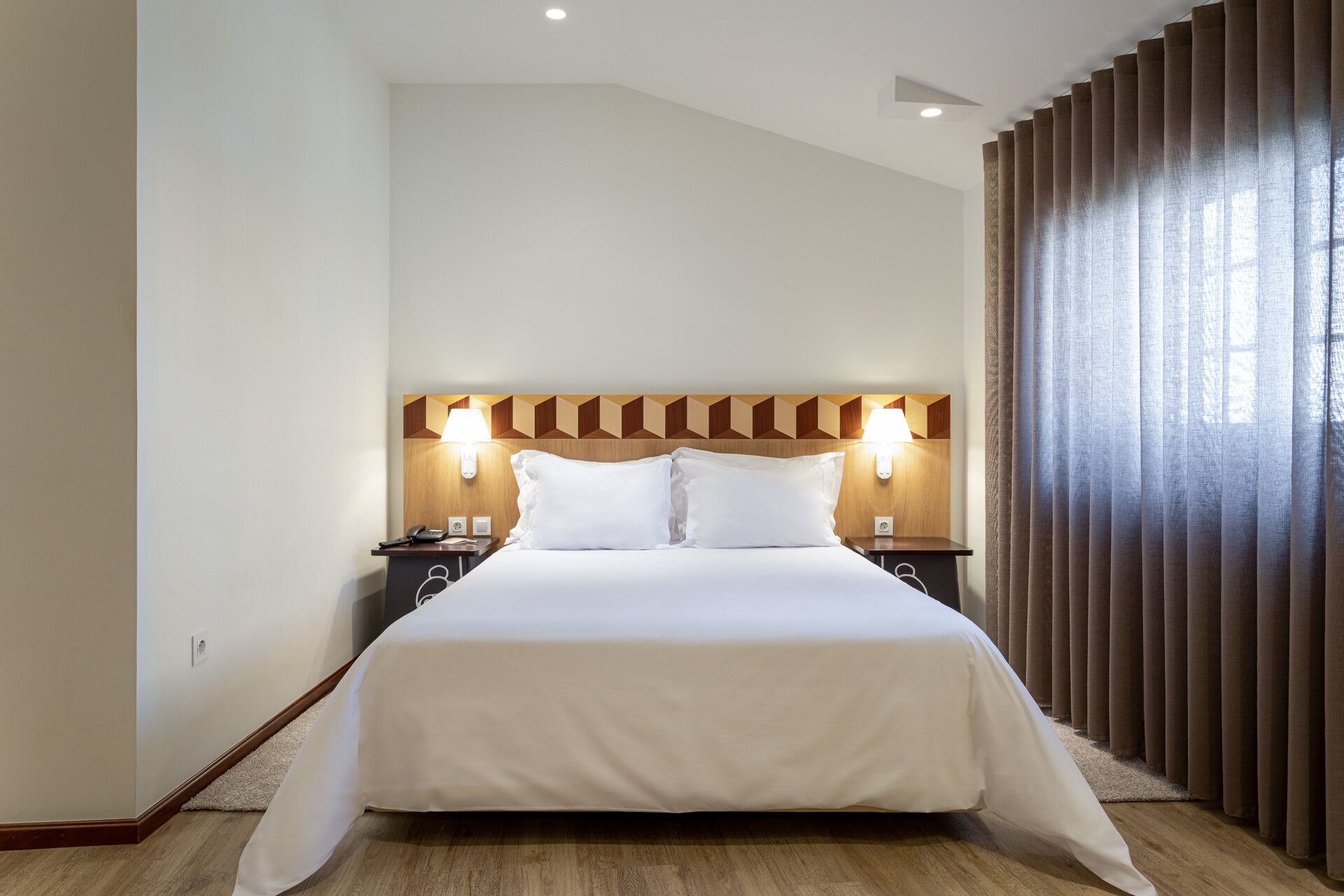 Una habitación de hotel con una cama grande, cabecero de madera, lámparas de noche y una ventana con cortinas.