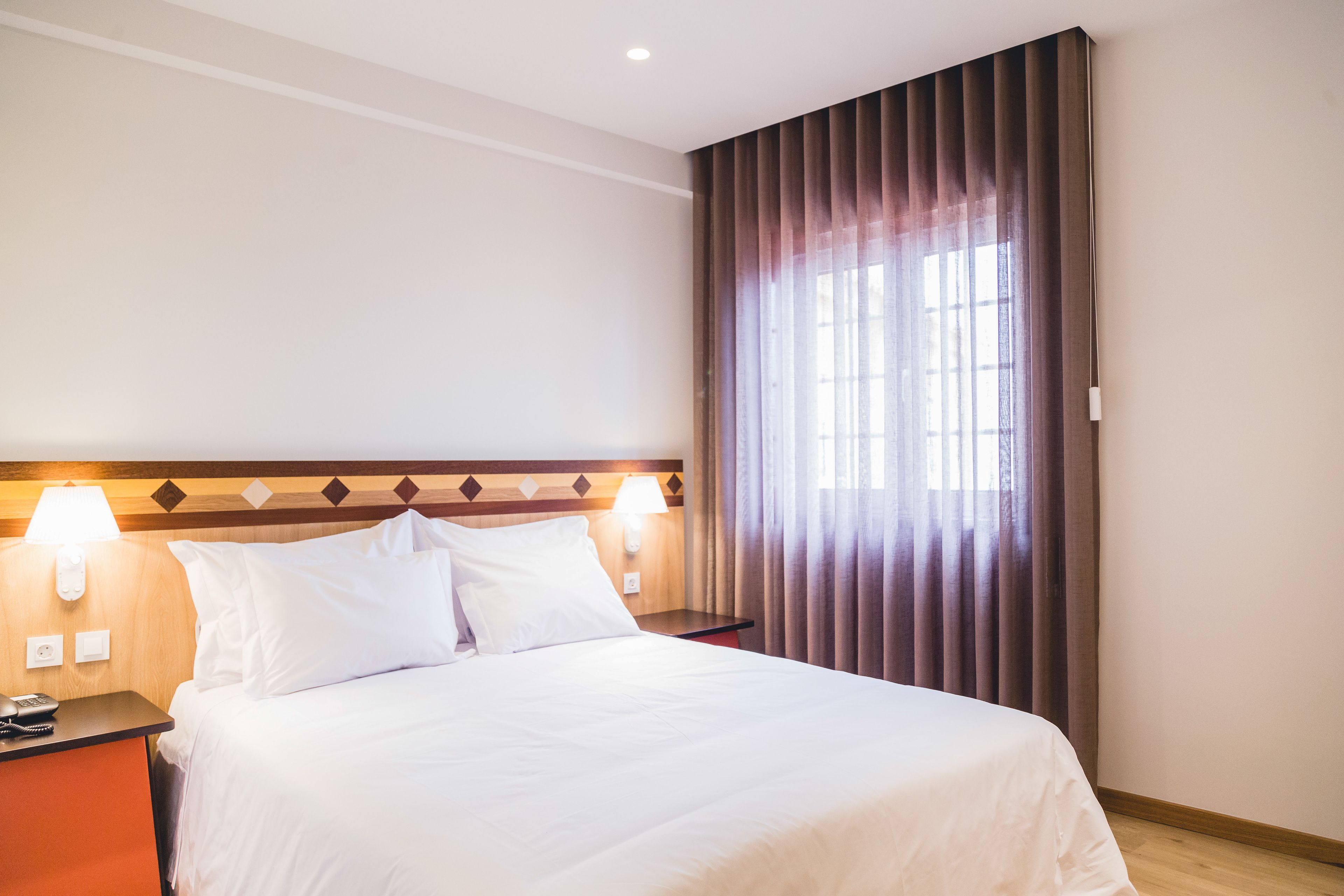A hotel room with a large bed, two bedside lamps, a nightstand with a phone, and a window with curtains.