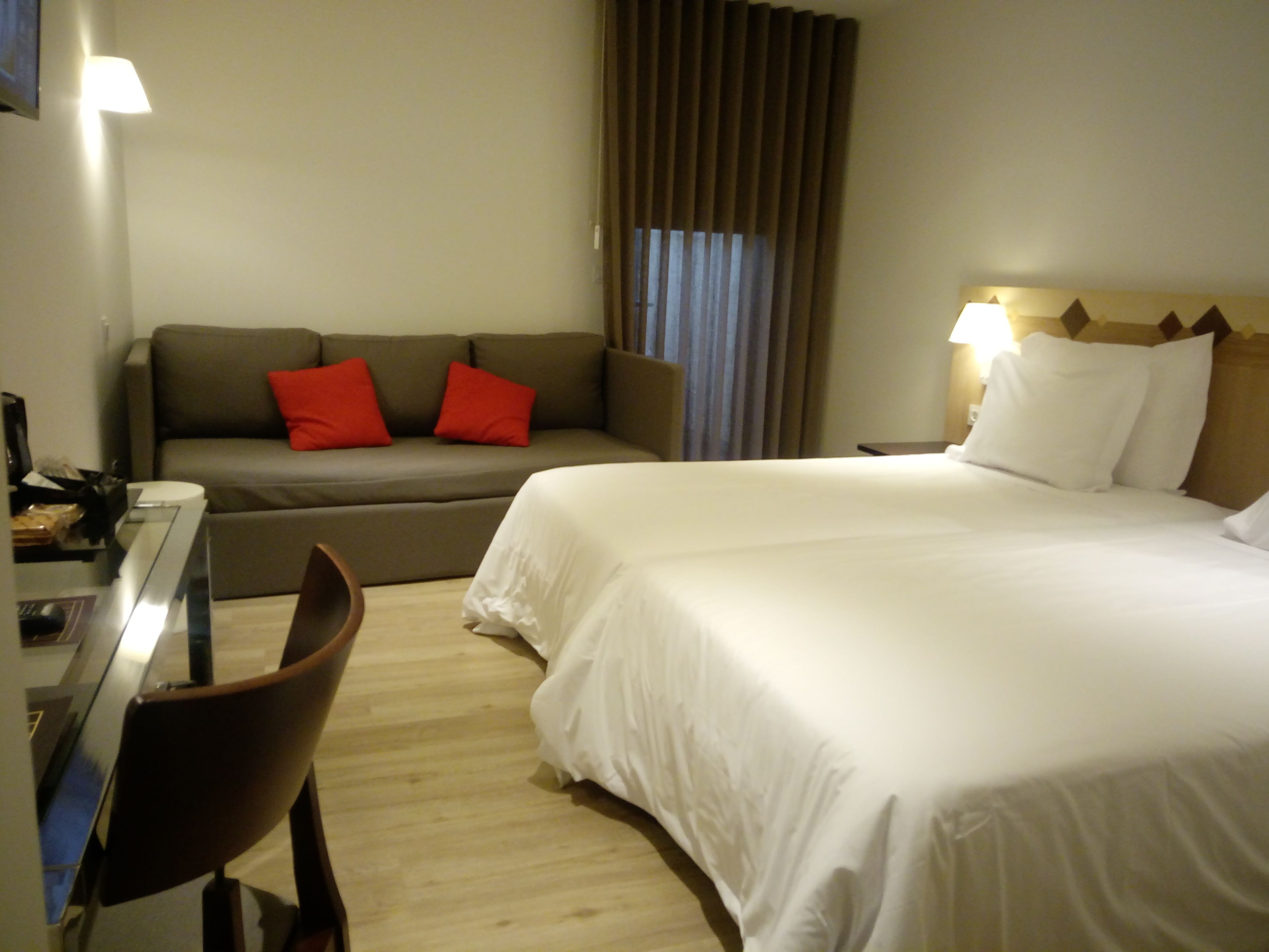 A hotel room with a bed, a sofa with red pillows, a desk with a computer, and a chair.