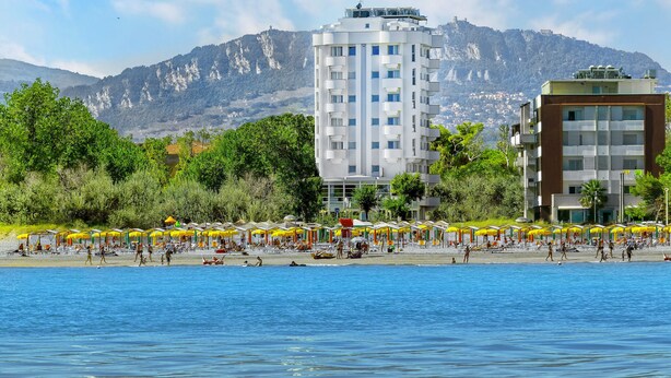 A coastal hotel with a poolside area, beach umbrellas, and a mountainous backdrop. - Hotel Meeting (Riccione)