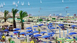 A beach with numerous blue umbrellas, palm trees, and sailboats on the water. - Hotel Meeting (Riccione)